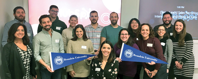 Fifteen smiling people, some holding blue Brandeis pennants, in a corporate space with large screens behind them.